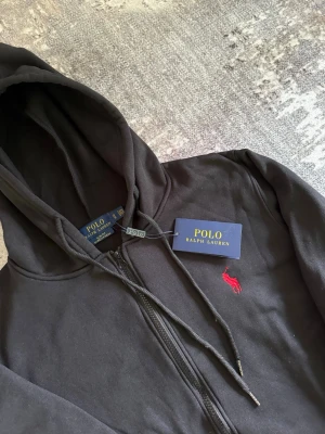 Svart hoodie från Polo Ralph Lauren - Svart hoodie med dragkedja från Polo Ralph Lauren. Klassisk design med huva, fickor framtill och justerbara snören. Ikoniska röda Polo-loggan broderad på bröstet. Tillverkad i mjukt bomullsmaterial för skön känsla.