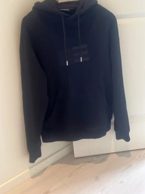 Lexington hoddie svart - Säljer en svart hoodie från Lexington med klassisk huva och dragsnören. På bröstet finns en diskret ton-i-ton logga. Hoodien har en avslappnad passform och är tillverkad i mjukt material, perfekt för en chill och stilren look. Storlek: S (passar M). Hör av dig vid frågor och funderingar