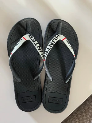 Svarta flip-flops från Hunter 39 - Stilrena svarta flip-flops från Hunter med vita remmar och svarta logotyper samt röda detaljer. Tillverkade i slitstark gummi med klassisk tårem och räfflad sula för extra grepp. Perfekta för sommaren och strandhäng.