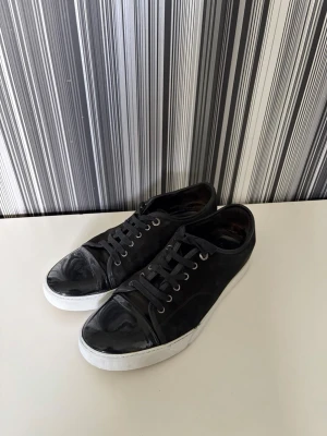 Svarta Lanvin - Snygga svarta sneakers med vit sula och lackad tådel. Skorna har klassisk snörning med svarta skosnören och metallöljetter. Ovandelen är i textil och tån är i glansigt material som ger en cool kontrast. Perfekta för en stilren och urban look.