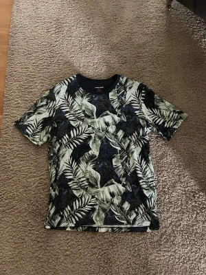 Mönstrad t-shirt från Jack & Jones - Snygg t-shirt från Jack & Jones med tropiskt bladmotiv i svart, grått och grönt. T-shirten har rund halsringning och korta ärmar, perfekt inför sommar säsongen. Den är helt oanvänd så den är som ny. Hör av er om ni har frågor!