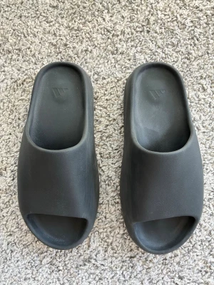 Adidas YZY Slide  - Säljer ett par mörkgråa Adidas YZY Slide i stilren och chunky design. Klassisk YZY-vibe med diskret logga i sulan. Sparsamt använda och bara använda inomhus, de är i mkt fint skick! Kan skickas i originalkartong!