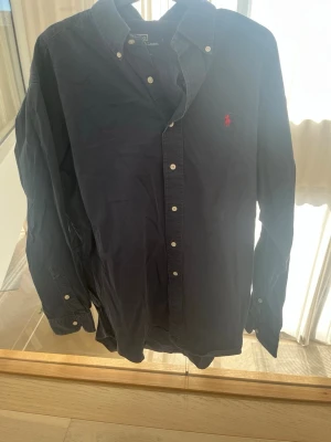Mörkblå skjorta från Polo Ralph Lauren - Klassisk mörkblå skjorta från Polo Ralph Lauren med röd broderad logga på bröstet. Skjortan är tillverkad i 100% bomull och har knappar framtill samt på ärmsluten. Perfekt för en stilren look.