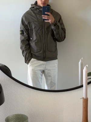 Stone Island grön vindjacka med huva - Snygg grön vindjacka från Stone Island med huva och dragkedja framtill, äkta såklart. Jackan har två stora fickor och den klassiska logotypen på vänster ärm. Knapparna på ena ärmen är skräddarsydda. Pris ej hugget i sten, skriv gärna vid intresse!