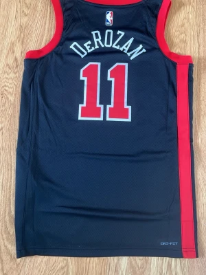 Chicago Bulls basketlinne DeRozan #11 - Snyggt basketlinne från Nike med Chicago Bulls-tryck och DeRozan #11. Linnet är svart med röda detaljer och har Dri-FIT-material för bra andningsförmåga. Perfekt för basketträning eller att bära som streetwear.