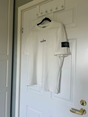 Vit Stone Island t-shirt med logga - Vit t-shirt från Stone Island med broderad logga på bröstet och den klassiska patchen på ärmen. T-shirten har en avslappnad passform och är tillverkad i mjuk bomull. Perfekt för en stilren och trendig look.