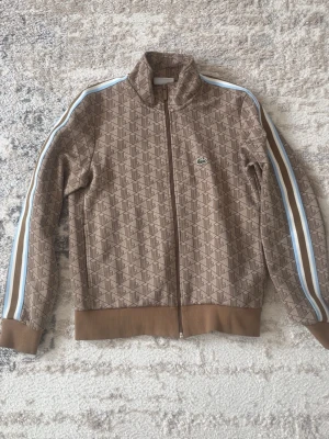 Brun mönstrad track jacket från Lacoste - Snygg brun track jacket från Lacoste med diskret mönster och klassisk krokodillogga på bröstet. Jackan har långa ärmar med blå och vita ränder längs sidorna samt dragkedja framtill. Perfekt för en sportig och trendig look.