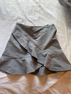 Grå volangkjol med brodyr - Säljer en snygg grå kjol med volangdetaljer och broderad texten 'Dreamer' längst ner. Kjolen har omlott-look och dragkedja i sidan. Perfekt för dig som gillar en trendig och stilren vibe.