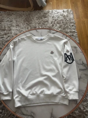 Vit sweatshirt från Moncler - Vit sweatshirt från Moncler med rund halsringning och ribbade muddar. Tröjan har Moncler-logga på bröstet och ett stort svart Moncler-emblem på ena ärmen. Tillverkad i mjuk bomull för en skön och avslappnad look.