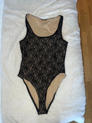 Beige body med svart spets - Snygg body i beige med svart spetsmönster över hela plagget. Ärmlös modell med rund halsringning och högt skurna ben. Perfekt för att skapa en trendig och elegant look.