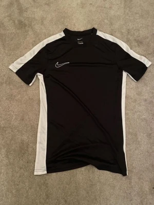 Svart Nike sporttröja med vita detaljer - Snygg svart sporttröja från Nike med vita paneler längs sidorna och axlarna. Tröjan har korta ärmar och är tillverkad i ett lätt och ventilerande material, perfekt för träning eller fotboll. Klassisk Nike-logga på bröstet.