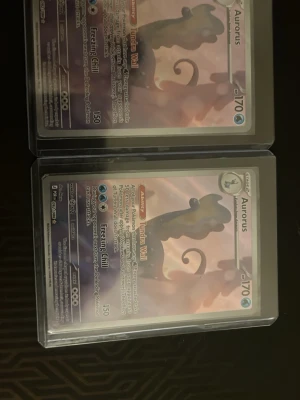Pokémonkort Aurorus x2 - Två stycken Pokémonkort med motivet Aurorus. Korten har en silvrig och lila design med detaljer i blått och vitt. Båda korten är inplastade i skyddsfodral. Perfekt för samlare eller Pokémonfans.