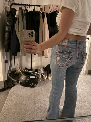 Blå bootcut jeans från True Religion - Säljer ett par ljusblå bootcut jeans från True Religion med ikoniska kontrastsömmar och detaljerade bakfickor med lock och knappar. Jeansen har låg midja och klassisk femficksdesign. Materialet är denim i bomull med en snygg tvätt.