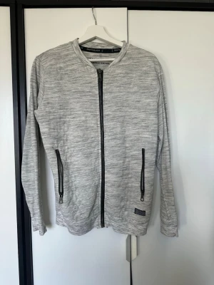 Ljusgrå ziptröja från Sam's Black - Ljusgrå ziptröja från Sam's Black med svarta dragkedjor framtill och på fickorna. Tröjan har en rak passform, rund halsringning och diskret mönster i tyget. Perfekt för en avslappnad stil.
