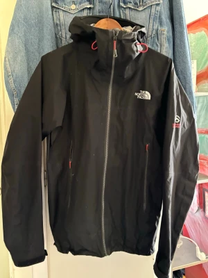 Svart regnjacka från The North Face - Svart regnjacka från The North Face med huva och hel dragkedja framtill. Jackan har röda detaljer på dragkedjor och snören, samt logga på bröstet och ärmen. Tillverkad i ett lätt och vattentätt material, perfekt för blöta dagar.
