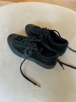 Svarta Converse One Star sneakers - Snygga svarta Converse One Star sneakers i mocka med klassisk stjärndetalj på sidan och svart sula. Skorna har lågt skaft, svarta skosnören och diskret All Star-logga på plösen. Perfekta för en stilren och avslappnad look.