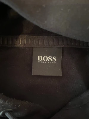 Svart hoodie från Hugo Boss - Svart hoodie från Hugo Boss med stor vit BOSS-logga på bröstet. Klassisk modell med huva och dragsko samt en stor magficka framtill. Perfekt för en stilren och avslappnad look.