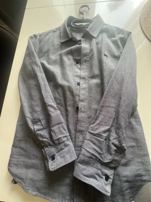 Grå skjorta från H&M med knappar - Snygg grå skjorta från H&M med klassisk krage och knäppning framtill. Skjortan har långa ärmar med manschett och en liten broderad detalj på bröstet. Perfekt för en stilren look.