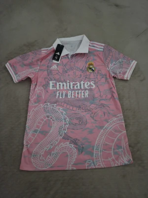 Rosa Real Madrid Adidas tröja - Säljer en rosa Real Madrid-tröja från Adidas med vit krage och drakmönster. Tröjan har klubbmärke på bröstet och 'Emirates Fly Better' tryckt framtill. Perfekt för dig som vill sticka ut på planen eller på läktaren.