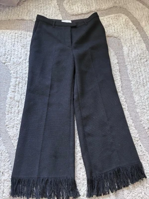 Svarta kostymbyxor med frans från H&M - Stilrena svarta kostymbyxor från H&M Edition med vida ben och coola fransdetaljer längst ner. Byxorna har hällor för skärp och klassisk midja. Materialet är strukturerat och ger en trendig känsla.