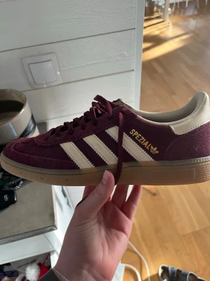 Adidas Spezial vinröda sneakers - Snygga Adidas Spezial sneakers i vinrött mocka med klassiska vita ränder och detaljer. Skon har gummisula och guldfärgad logga på sidan. Låg modell med retrovibbar och bekväm passform. Perfekt för dig som gillar streetstyle och sportig look.