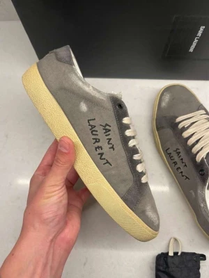 Gråa Saint Laurent sneakers - Gråa sneakers från Saint Laurent med handskriven logga på sidan. Skorna har en vintageinspirerad look med slitna detaljer, vita snören och beige sula. Tillverkade i mocka och läder för en lyxig känsla och stilren design.