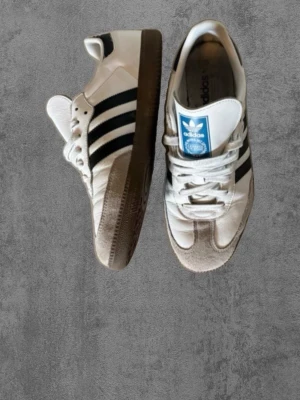 Adidas Samba OG vita sneakers - Klassiska Adidas Samba OG sneakers i vitt läder med svarta ränder och brun gummisula. Skorna har lågt skaft, snörning och blå detalj på plösen. Perfekta för en sportig och stilren look.