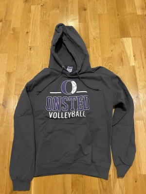 Mörkgrå Jerzees Onsted Volleyball hoodie - Snygg mörkgrå hoodie från Gildan med tryck i lila och vitt där det står 'Onsted Volleyball' och en volleyboll ovanför texten. Klassisk känguruficka framtill och justerbar huva med dragsko. Perfekt för dig som gillar sportig stil.
