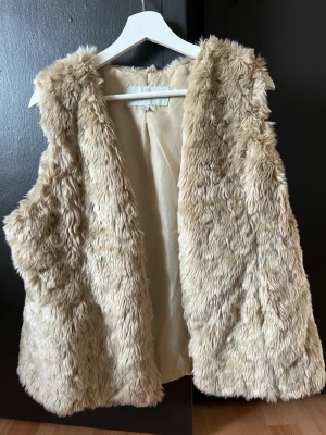Beige fluffig pälsväst - Snygg beige pälsväst i fuskpäls med mjukt foder. Västen har en rak passform och är ärmlös, perfekt för att ge din outfit en trendig touch. Passar till många olika stilar och är superenkel att matcha med andra plagg. Lämna gärna prisförslag. 