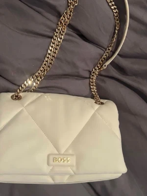 Vit axelväska från BOSS med kedja - Snygg vit axelväska från BOSS med quiltat mönster och guldfärgad kedjerem. Väskan har en stilren design med BOSS-logga framtill och passar perfekt för dig som gillar en clean och modern look.