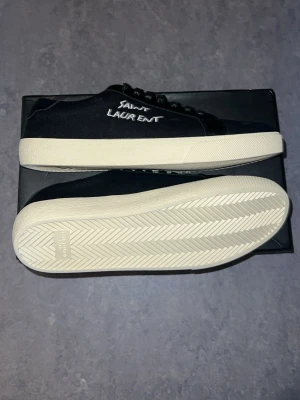 Svarta Saint Laurent sneakers - Svarta sneakers från Saint Laurent med vit broderad logga på sidan och vit sula. Skorna har klassisk låg profil, svarta skosnören och ovandel i canvas och läder. Snygg och stilren design med exklusiv känsla.