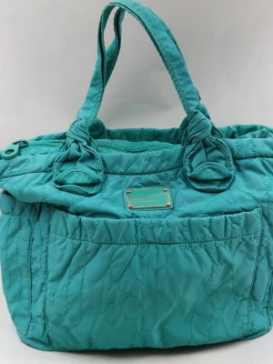 Turkos handväska Marc by Marc Jacobs - Snygg turkos handväska från Marc by Marc Jacobs med quiltad design och dubbla handtag med knutdetaljer. Väskan har en metallbricka framtill och är tillverkad i nylonmaterial. Perfekt för dig som vill ha en trendig och rymlig väska med cool workwear-vibe. Den långa remmen saknas men annars fint begagnat skick.