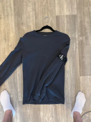 Mörkgrå långärmad tröja Calvin Klein - Snygg mörkgrå långärmad tröja från Calvin Klein med rund halsringning och diskret CK-logga på ärmen. Tröjan har en ribbad struktur och är perfekt för en stilren och enkel look. Väldigt bra skick, aldrig andvänd. Storlek S. 