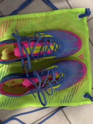Adidas F50  - Säljer ett par Adidas F50 fotbollsskor i en riktigt snygg färgkombo av blått, rosa och neongult. Skorna har snörning, låg profil och är tillverkade i syntetmaterial för lätt vikt och bra bollkontroll. Perfekta för dig som vill sticka ut på planen!