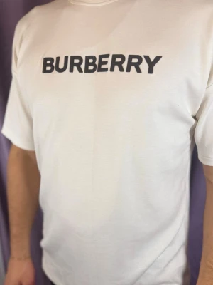 Vit Burberry t-shirt med logga - Vit t-shirt från Burberry med klassisk svart logotyp tryckt över bröstet. T-shirten har en rak passform och korta ärmar. Perfekt för dig som gillar stilrena och exklusiva plagg. Materialet känns mjukt och skönt mot huden.
