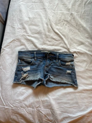 Blå slitna jeansshorts - Säljer ett par blå jeansshorts med slitna detaljer och fransig kant. Klassisk femficksmodell med låg midja och råa kanter för en avslappnad look. Perfekt för varma dagar och festivaler.