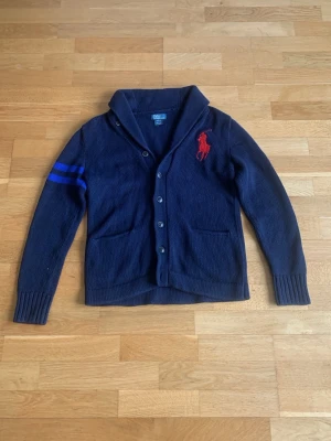 Mörkblå kofta från Polo Ralph Lauren - Mörkblå stickad och stilren kofta från Polo Ralph Lauren. Stora röda loggan på bröstet och blå ränder samt röd siffra 3 på ärmen. Knäpps med knappar framtill och har två fickor. Perfekt för en stilren look.