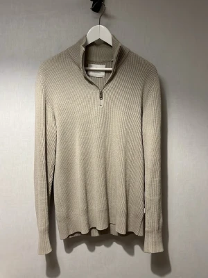 Beige stickad tröja med halv dragkedja - Snygg beige stickad tröja med ribbad struktur och hög krage med halv dragkedja. Långärmad modell som passar perfekt till lager på lager. Tröjan har en stilren och minimalistisk look, vilket gör den enkel att matcha med olika outfits.