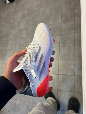 Vita Adidas fotbollsskor med röd detalj - Säljer ett par snygga vita Adidas fotbollsskor med svarta ränder och röda detaljer vid hälen. Skorna har snörning, låg profil och en ovandel i syntetmaterial med ventilerande mesh. Sulan har dobbar för bra grepp på planen. Perfekta för dig som vill sticka ut på matchen!