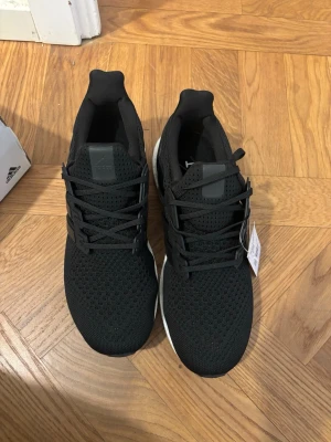 Svarta Adidas Ultraboost sneakers - Svarta Adidas Ultraboost sneakers med stickad ovandel i mesh och klassiska tre ränder på sidan. Skorna har en vit Boost-sula för extra komfort och svart yttersula. Snygg och sportig design med snörning och låg profil.