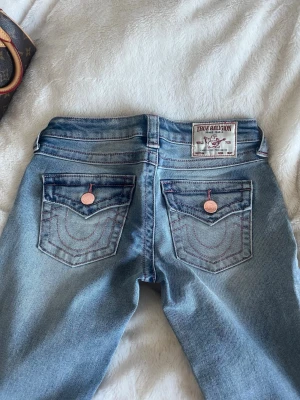 True Religion ljusblå jeans - Säljer ett par snygga ljusblå jeans från True Religion med ikoniska bakfickor och kontrastsömmar i rött. Jeansen har klassisk femficksdesign, kopparfärgade knappar och patch med logga bak i midjan. Perfekta för en cool och avslappnad stil.