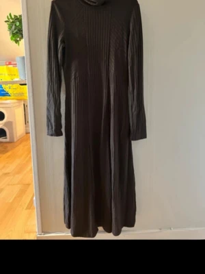 Mörkbrun långärmad klänning med polokrage - En stilren mörkbrun långklänning med lång ärm och polokrage. Klänningen har en ribbad struktur framtill som ger en snygg detalj. Perfekt för dig som gillar en enkel men elegant look.