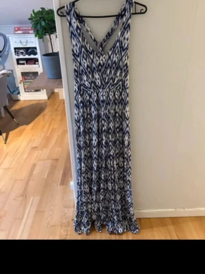 Lång blåvit mönstrad klänning - Säljer en långklänning med blått och vitt grafiskt mönster. Klänningen har v-ringning och breda axelband som korsas i ryggen. Materialet känns mjukt och svalt, perfekt för varma dagar. Passformen är normal och modellen är luftig och flowy.