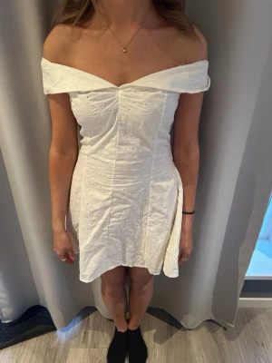 Vit offshoulder kortklänning - Söt vit kortklänning med offshoulder-design och broderade detaljer. Klänningen har en figurnära passform upptill, smockad rygg och lätt utställd kjol. Perfekt för sommarens alla tillfällen.