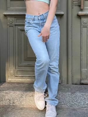 Levis 501 - Säljer ett par ljusblå jeans med rak passform och hög midja. Klassisk femficksmodell med bälteshällor och knappgylf. Jeansen har en avslappnad look och är perfekta till sneakers. Materialet är denim i bomull. Allt i dm