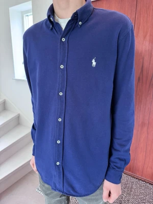 Blå skjorta från Ralph Lauren - Stilren marinblå långärmad tröja med knappar i halsen. Mycket mjukt och skönt material. Perfekt som ett lite mer dressat alternativ till en vanlig t-shirt eller piké.  • Märke: Polo Ralph Lauren • Storlek: Large men passar Medium (Slim Fit) • Skick: 9/10 (inga slitage)  • Färg: Marinblå  
