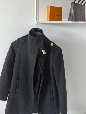 Svart kappa med guldknappar - Stilren svart kappa med hög krage och dekorativa guldknappar på axel och krage. Kappan har raka fickor och en klassisk, rak passform. Perfekt för dig som vill ha en elegant och tidlös look till höst och vinter.