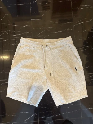 Grå shorts från Polo Ralph Lauren - Snygga ljusgrå shorts från Polo Ralph Lauren med klassisk broderad logga på vänster ben. Shortsen har snörning i midjan och två sidofickor. Perfekta för sommaren och riktigt sköna tack vare mjukt bomullsmaterial.
