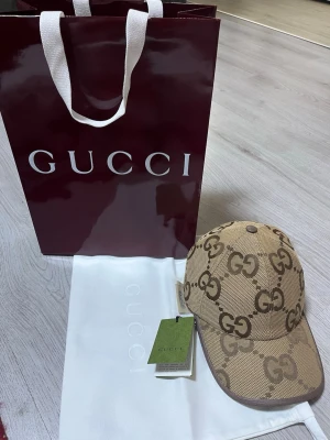 Beige keps från Gucci med GG-mönster - Säljer en beige keps från Gucci med det klassiska GG-monogrammet i brunt över hela kepsen. Kepsen har böjd skärm och justerbar rem bak. Tillverkad i canvasmaterial med läderdetaljer. Perfekt för dig som vill ha en lyxig och trendig accessoar. Kepsen storlek M, De som följer med är box, låda, tags, dust bag, 