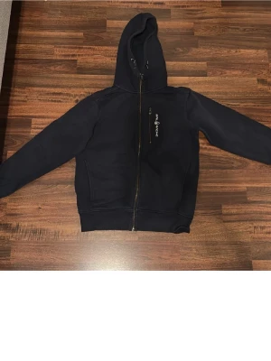 Marinblå hoodie från Sail Racing - Snygg marinblå hoodie från Sail Racing med dragkedja framtill och logga på bröstet. Klassisk huva med snörning och ribbade muddar vid ärmslut och nederkant. Perfekt för en avslappnad och sportig stil. Tillverkad i mjukt och bekvämt material.